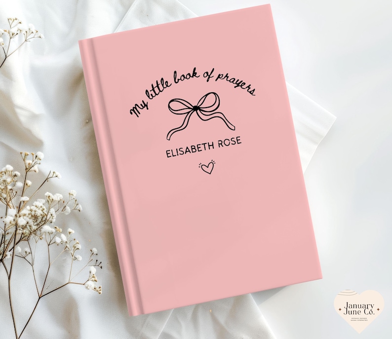 May include: A pink book with the text "my little book of prayers" in a handwritten font. Below the text is a bow illustration, the name "ELISABETH ROSE", and a small heart. The book is on a white surface.