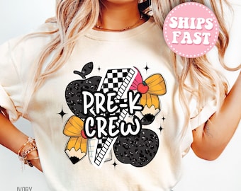 May include: Ivory-colored t-shirt featuring a colorful graphic design. The design includes the text "PRE-K CREW" with illustrations of an apple, pencil, ruler, and bow. The shirt has a round neck and short sleeves.