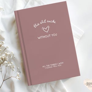 May include: A dusky pink journal with the text "this shit sucks without you" and "all the things I wish I could tell you" in white. A white heart symbol is between the first two lines of text. The journal is on a white surface.