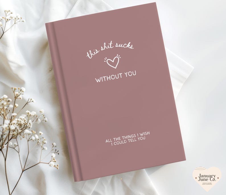 May include: A dusky pink journal with the text "this shit sucks without you" and "all the things I wish I could tell you" in white. A white heart symbol is between the first two lines of text. The journal is on a white surface.