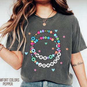 Puede incluir: Camiseta Comfort Colors gris oscuro con un dise&ntilde;o de collar de cuentas de colores. El collar deletrea "MRS JOHNSON" con corazones y estrellas. La camiseta tambi&eacute;n tiene las palabras "COMFORT COLORS PEPPER" impresas en la parte inferior.