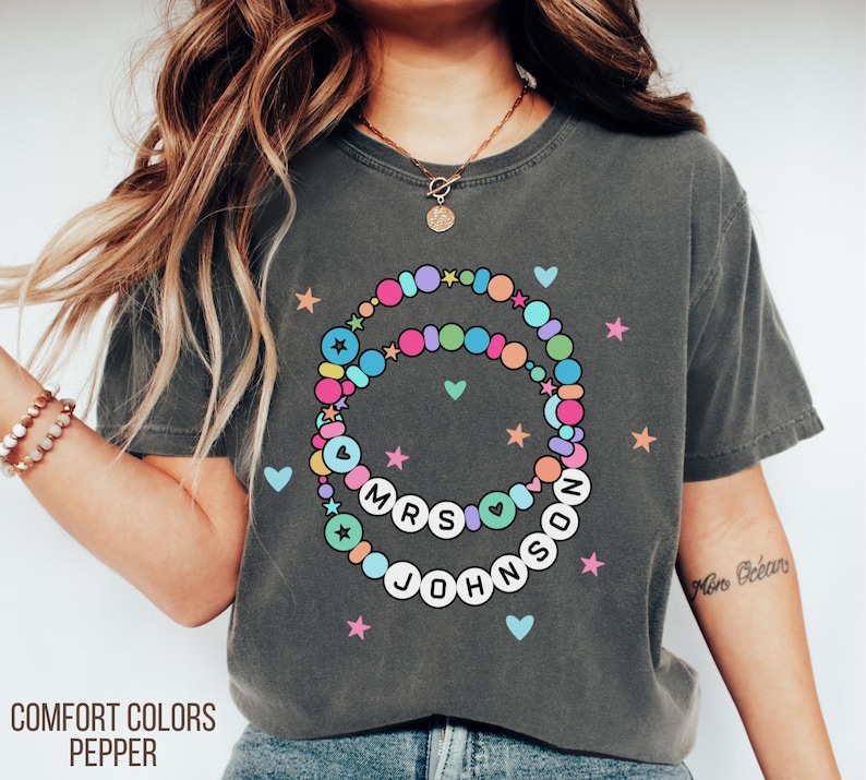 Puede incluir: Camiseta Comfort Colors gris oscuro con un dise&ntilde;o de collar de cuentas de colores. El collar deletrea "MRS JOHNSON" con corazones y estrellas. La camiseta tambi&eacute;n tiene las palabras "COMFORT COLORS PEPPER" impresas en la parte inferior.