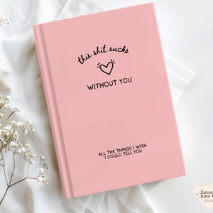 May include: A pink journal with the text "this shit sucks without you" and "all the things I wish I could tell you." The cover features a black heart graphic. The journal is on a white surface with floral accents.
