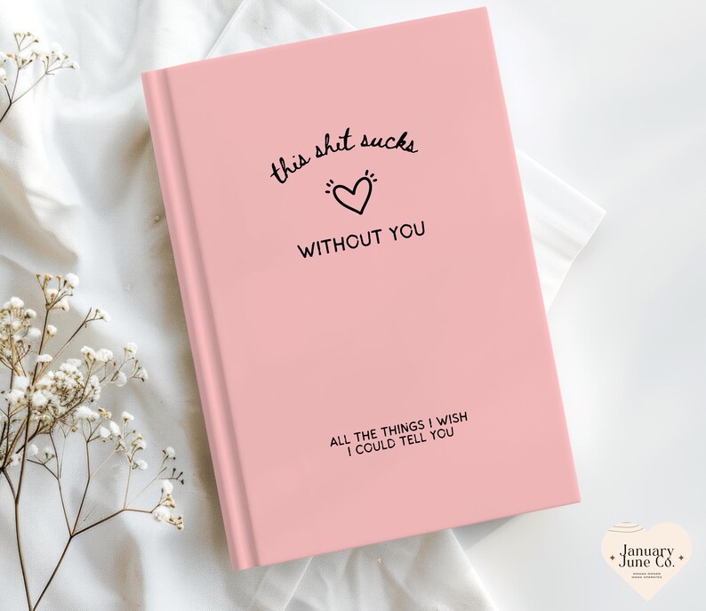 May include: A pink journal with the text "this shit sucks without you" and "all the things I wish I could tell you." The cover features a black heart graphic. The journal is on a white surface with floral accents.