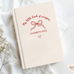 May include: A cream-colored book with the title "my little book of prayers" in red script, along with a bow illustration and the name "Elisabeth Rose." A small red heart is below the name. The book is on a white surface.
