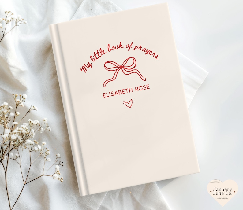 May include: A cream-colored book with the title "my little book of prayers" in red script, along with a bow illustration and the name "Elisabeth Rose." A small red heart is below the name. The book is on a white surface.