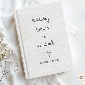 May include: A hardcover book with a textured, off-white cover. The cover features the handwritten text "birthday letters to michael ray" and the date "NOVEMBER 22, 2025" in black. The book is set against a white fabric background.