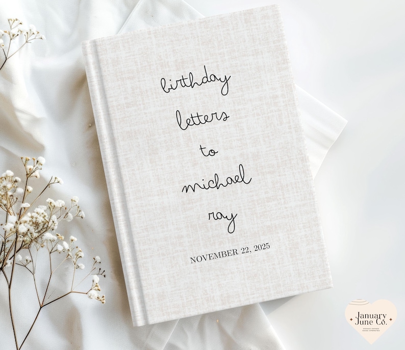 May include: A hardcover book with a textured, off-white cover. The cover features the handwritten text "birthday letters to michael ray" and the date "NOVEMBER 22, 2025" in black. The book is set against a white fabric background.