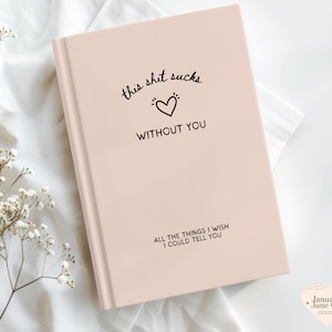 May include: A pale pink journal with the text "this shit sucks without you" and "all the things I wish I could tell you" in black script. A black heart is between the phrases. The journal is on a white surface with white flowers.