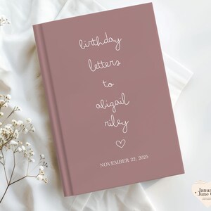 May include: A mauve-colored journal with the words "birthday letters to abigail riley" in white script. A heart symbol and the date "November 22, 2025" are also printed on the cover. The journal is on a white fabric surface.