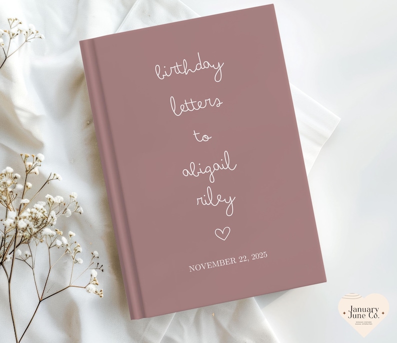 May include: A mauve-colored journal with the words "birthday letters to abigail riley" in white script. A heart symbol and the date "November 22, 2025" are also printed on the cover. The journal is on a white fabric surface.
