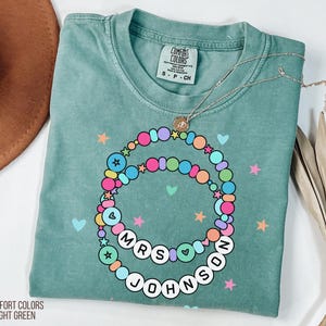 Puede incluir: Camiseta verde claro Comfort Colors con un dise&ntilde;o de collar de cuentas de colores. El collar deletrea "MRS JOHNSON" y est&aacute; adornado con estrellas y corazones. Tambi&eacute;n es visible un delicado collar de plata con un colgante.