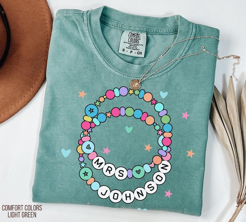 Puede incluir: Camiseta verde claro Comfort Colors con un dise&ntilde;o de collar de cuentas de colores. El collar deletrea "MRS JOHNSON" y est&aacute; adornado con estrellas y corazones. Tambi&eacute;n es visible un delicado collar de plata con un colgante.
