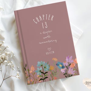 May include: A hardcover book with a dusty rose cover featuring the text "CHAPTER 13" and "a chapter worth remembering." The book is decorated with a floral border of blue, purple, and orange flowers. The name "Kaitlyn" is below a heart.