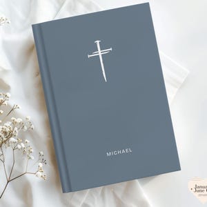 May include: A blue-grey book with a white cross and nail design. The name "MICHAEL" is printed in white at the bottom. The book is on a white surface with white flowers.