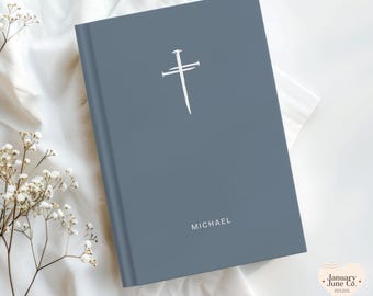 May include: A blue-grey book with a white cross and nail design. The name "MICHAEL" is printed in white at the bottom. The book is on a white surface with white flowers.