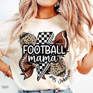 May include: Ivory-colored t-shirt with a football-themed graphic. The design includes footballs, a bow, and the text "FOOTBALL mama" in black. The footballs have a leopard print pattern. The shirt also has the text "#45 Johnson".