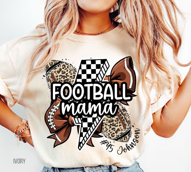 May include: Ivory-colored t-shirt with a football-themed graphic. The design includes footballs, a bow, and the text "FOOTBALL mama" in black. The footballs have a leopard print pattern. The shirt also has the text "#45 Johnson".