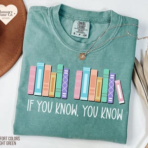 May include: Light green Comfort Colors t-shirt with a colorful bookshelf graphic and the text "IF YOU KNOW, YOU KNOW." The shirt has a crew neck and short sleeves. A silver necklace with a pendant is on the shirt.