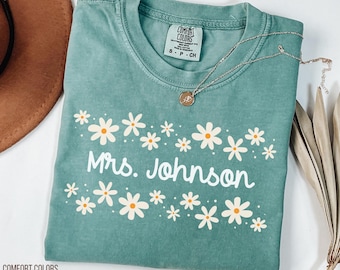 May include: Light green Comfort Colors t-shirt with the name "Mrs. Johnson" in white script, surrounded by a daisy floral design. A silver necklace with a round pendant is on the shirt.