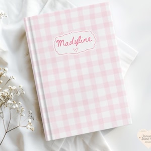 May include: A pink and white gingham patterned journal with the name "Madlyine" in pink script on the cover. The journal has a heart symbol below the name. The journal is on a white surface with a sprig of white flowers.