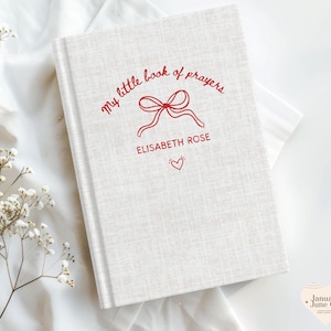 May include: A hardcover book with a light beige linen cover. The title "my little book of prayers" is written in red script, along with the name "ELISABETH ROSE" and a heart icon. The book is on a white fabric surface.