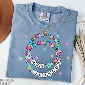 Puede incluir: Camiseta azul vaquero Comfort Colors con un dise&ntilde;o de collar de cuentas de colores. El collar tiene el texto "MRS JOHNSON" en letras blancas. La camiseta tambi&eacute;n presenta estrellas y corazones.