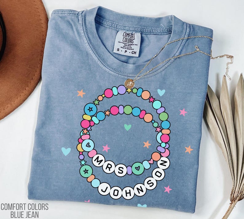 Puede incluir: Camiseta azul vaquero Comfort Colors con un dise&ntilde;o de collar de cuentas de colores. El collar tiene el texto "MRS JOHNSON" en letras blancas. La camiseta tambi&eacute;n presenta estrellas y corazones.