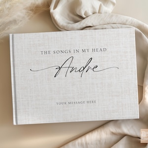 May include: A rectangular hardcover book with a light gray, textured fabric cover. The front cover features the black printed text THE SONGS IN MY HEAD, the name Andre in a large black script font, and the text YOUR MESSAGE HERE below it.