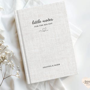 May include: A hardcover notebook with the text "little notes for the big day" and the names "Amanda & Mark" printed in black. The notebook has a textured, off-white cover and a heart-shaped design. The notebook is on a white surface.