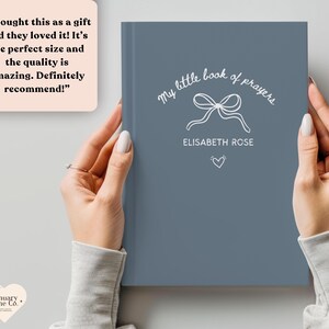 May include: A blue book titled "My Little Book of Prayers" with white script and a bow illustration. The book is held by a person. The name "Elisabeth Rose" is printed below the bow, with a heart symbol beneath it.
