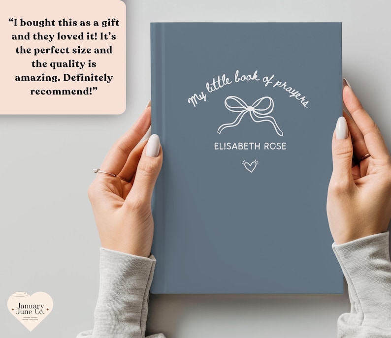 May include: A blue book titled "My Little Book of Prayers" with white script and a bow illustration. The book is held by a person. The name "Elisabeth Rose" is printed below the bow, with a heart symbol beneath it.