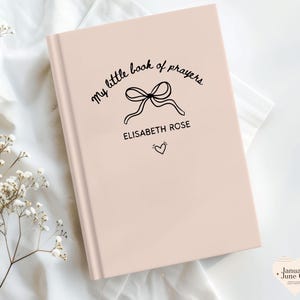 May include: A light pink book with the text "my little book of prayers" in a cursive font. A bow illustration is centered above the name "ELISABETH ROSE" and a small heart. The book is on a white surface.