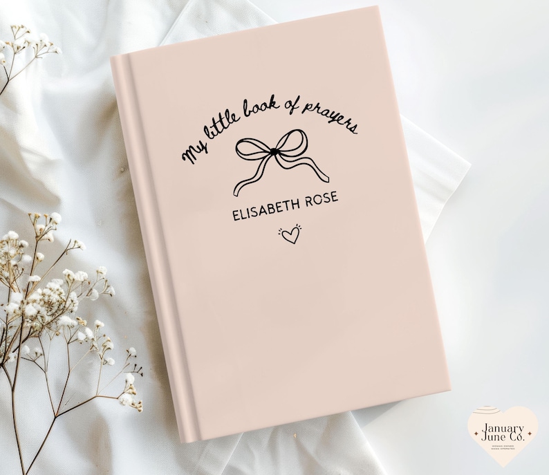 May include: A light pink book with the text "my little book of prayers" in a cursive font. A bow illustration is centered above the name "ELISABETH ROSE" and a small heart. The book is on a white surface.