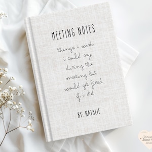 May include: A hardcover notebook with the text "MEETING NOTES" and humorous phrases about meetings. The notebook is light beige with black text and the name "NATALIE" at the bottom. The notebook is on a white surface.