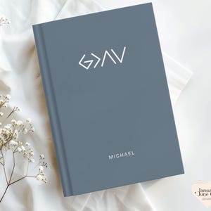 May include: A slate blue journal with the text "G>AV" and the name "MICHAEL" in white. The journal is closed and resting on a white cloth, with sprigs of white flowers in the corner.