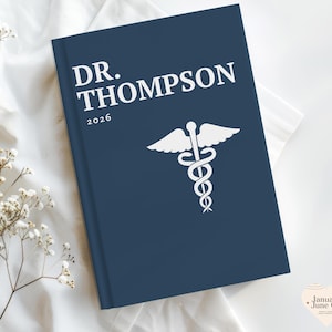 May include: A dark blue hardcover journal featuring the white text DR. THOMPSON and the year 2026 printed on the front cover. Below the text is a white graphic of a caduceus symbol, which consists of a winged staff with two snakes coiled around it.