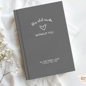 May include: A grey journal with the text "this shit sucks without you" and "all the things I wish I could tell you" in white. A white heart graphic is between the phrases. The journal is on a white surface.