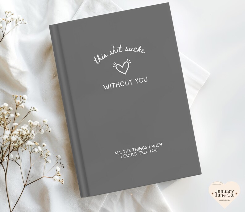 May include: A grey journal with the text "this shit sucks without you" and "all the things I wish I could tell you" in white. A white heart graphic is between the phrases. The journal is on a white surface.