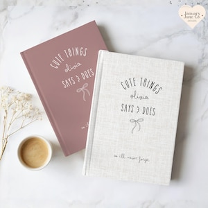 May include: Two hardcover journals with the text "CUTE THINGS olivia SAYS & DOES" and a bow illustration. One journal is dusty rose, the other is off-white. A small cup of coffee and sprigs of white flowers are also in the image.