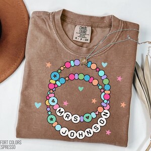 Puede incluir: Una camiseta marr&oacute;n Comfort Colors Espresso con un dise&ntilde;o de collar de cuentas de colores. El collar deletrea "MRS JOHNSON" en cuentas de letras blancas. La camisa tambi&eacute;n presenta corazones y estrellas.