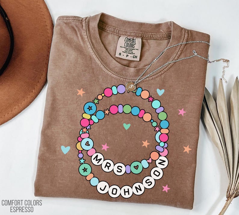 Puede incluir: Una camiseta marr&oacute;n Comfort Colors Espresso con un dise&ntilde;o de collar de cuentas de colores. El collar deletrea "MRS JOHNSON" en cuentas de letras blancas. La camisa tambi&eacute;n presenta corazones y estrellas.