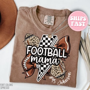 May include: A brown Comfort Colors Espresso t-shirt featuring a graphic that says "FOOTBALL mama" with a lightning bolt, footballs, and a bow. The shirt has a crew neck and short sleeves. The graphic includes the number 45 and the name Johnson.