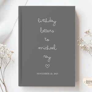 May include: A gray journal with the words "birthday letters to michael ray" in white script. A heart symbol and the date "NOVEMBER 22, 2025" are also included. The journal is surrounded by white flowers.