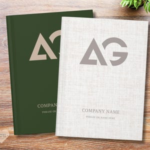 May include: Two hardcover notebooks, one dark green and one off-white, with the letters "AG" in a minimalist design. The green notebook has a beige logo, and the off-white notebook has a gray logo. Both have the text "COMPANY NAME" and "PHRASE OR NAME HERE".