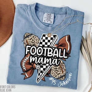 May include: Blue jean Comfort Colors t-shirt with a graphic that says "Football Mama". The graphic includes a football, a bow, and a lightning bolt. A gold necklace is draped across the shirt.