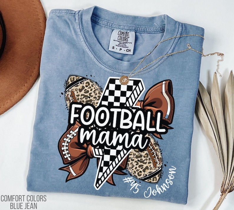 May include: Blue jean Comfort Colors t-shirt with a graphic that says "Football Mama". The graphic includes a football, a bow, and a lightning bolt. A gold necklace is draped across the shirt.