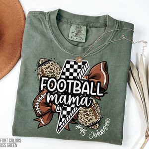 May include: A moss green t-shirt with a graphic that says "Football Mama". The graphic includes a lightning bolt, footballs with leopard print, and a brown bow. The shirt also has the name "Johnson" and the number "45".