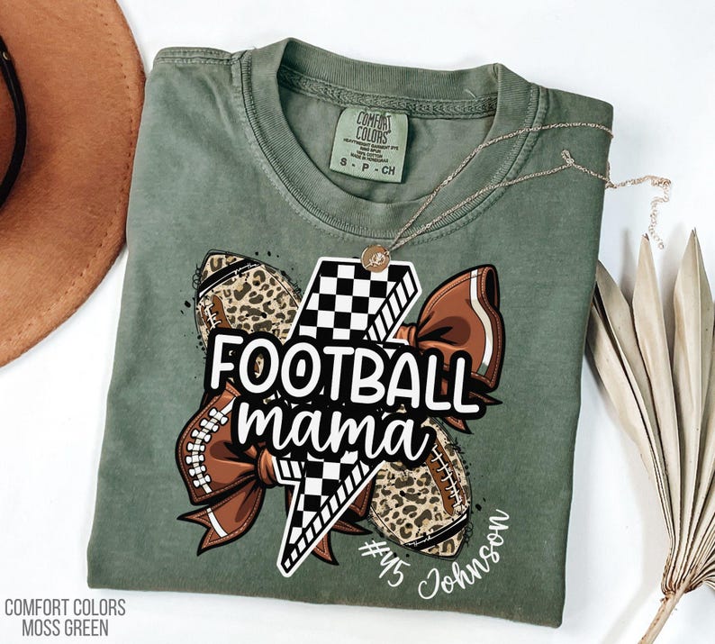 May include: A moss green t-shirt with a graphic that says "Football Mama". The graphic includes a lightning bolt, footballs with leopard print, and a brown bow. The shirt also has the name "Johnson" and the number "45".