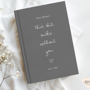 Peut inclure: Un journal gris avec le texte "Dear Michael, this shit sucks without you, Love, Amy" en &eacute;criture blanche. Un petit c&oelig;ur blanc est en dessous du texte. Le journal est sur une surface blanche avec des fleurs blanches.